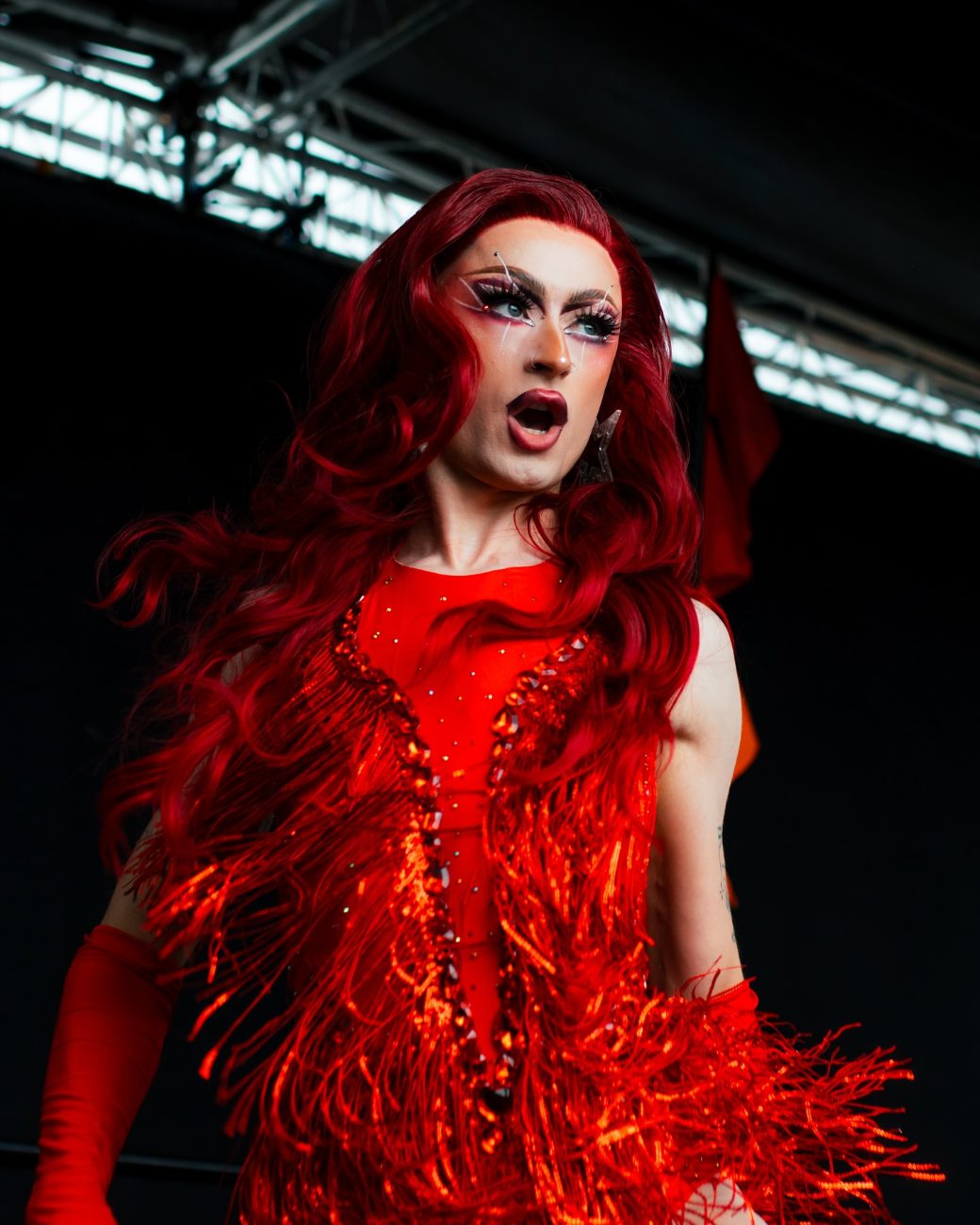 Charlie Sin in a red wig and red fringed sequin outfit, performing on an outdoor stage