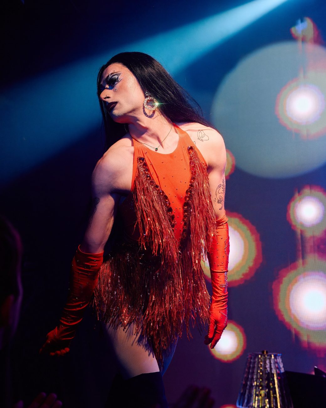 Charlie Sin in an orange fringed bodysuit and long gloves under blue stage light