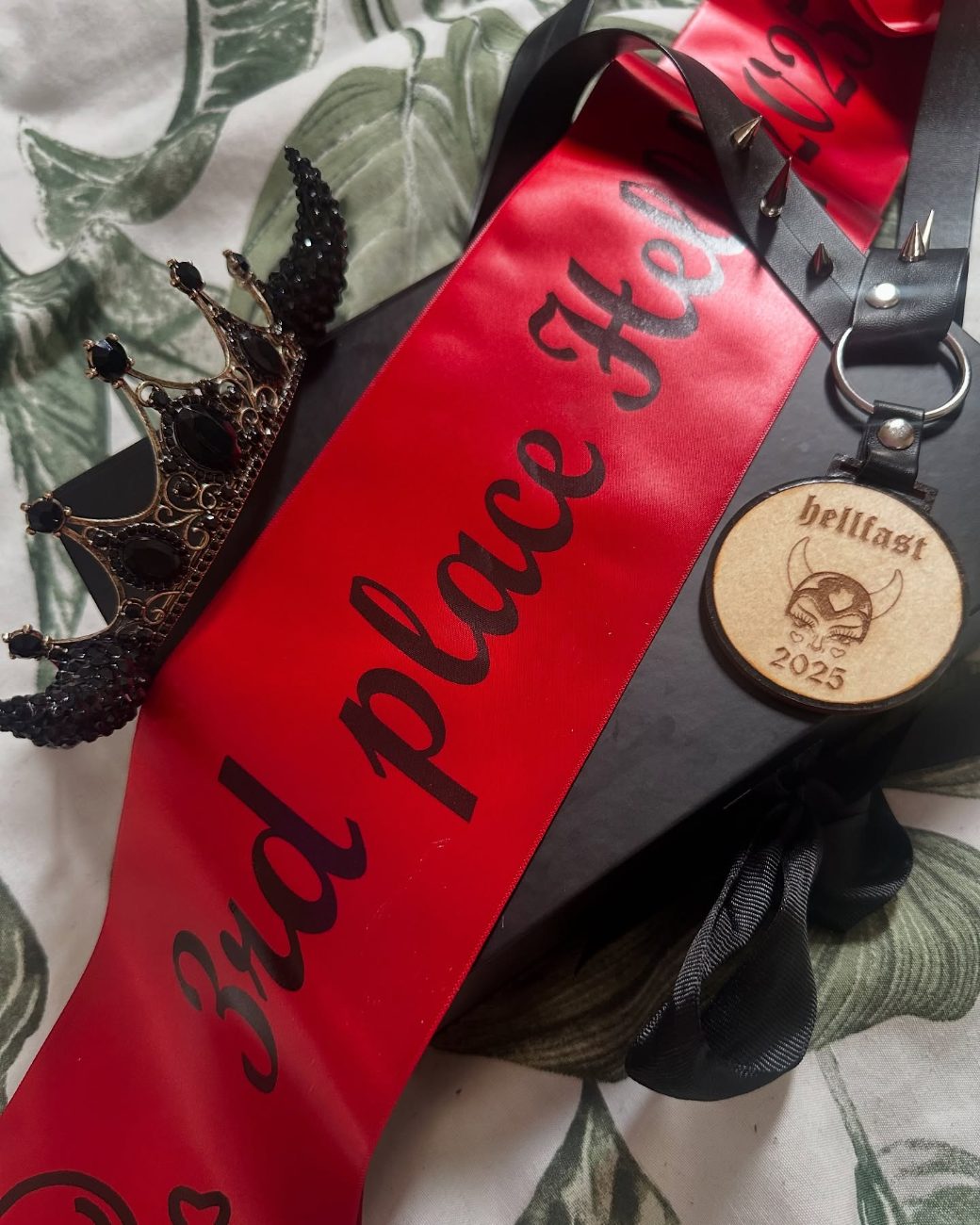 A red third place sash, a black crown and a Hellfast 2025 medal